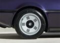 A close-up view of a silver alloy wheel on a dark purple BMW M1, showcasing a unique design with multiple circular slots.