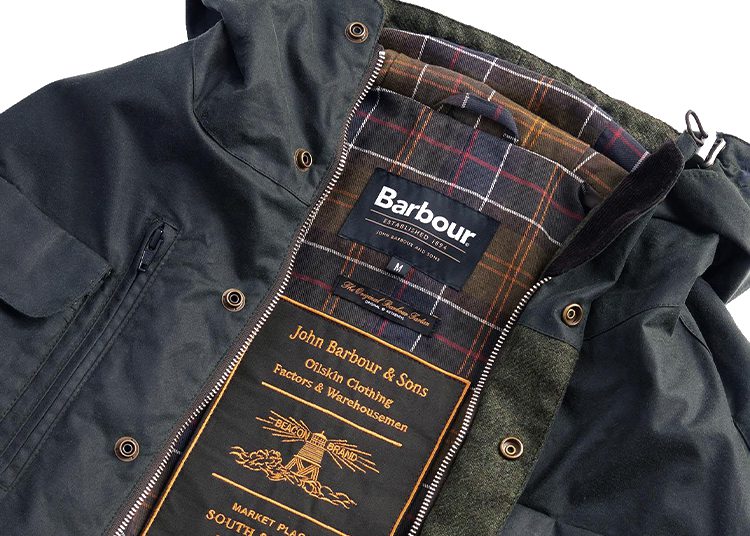 Close-up of a Barbour jacket from the 2024 Holiday Gift Guide, showcasing its interior plaid lining and iconic brand label. The label reads "John Barbour & Sons" with additional company details, making it a refined choice for any duPont REGISTRY enthusiast.