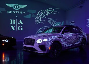 In a dimly lit showroom, a white Bentley Bentayga with color-changing illuminated patterns takes center stage, surrounded by branding and abstract designs on the walls.