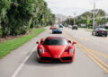 Red sports car driving on a city street, followed by a blue car. Trees and buildings line the road, with other vehicles in adjacent lanes.