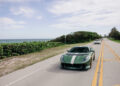 A green sports car with racing stripes drives on a coastal road, followed by another car. Lush greenery and the ocean line the roadside under a partly cloudy sky.