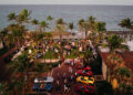 Aerial view of a seaside event with people gathered on a lawn surrounded by palm trees and luxury cars parked nearby.