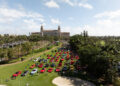 Aerial view of a car show on a grassy area with numerous cars displayed, surrounded by trees and pathways, with a large historic building in the background.