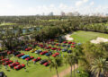 Aerial view of a car show on a grassy field, featuring numerous vintage cars and people. Palm trees and a golf course are in the background, with a city skyline in the distance.