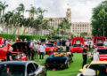 A car show featuring numerous high-end sports cars, including convertibles and coupes, with many attendees. Palm trees and a large building are in the background.