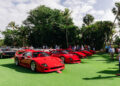 A row of red sports cars displayed on a grassy area, surrounded by people and trees under a partly cloudy sky.