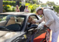 A man in a white suit and hat shakes hands with a person sitting in a black Ferrari on a red carpet. Passersby watch in the background.