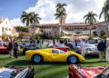 A gathering of people at a car show on a lawn, with vintage and modern cars on display. The background features an ornate building and palm trees under a partly cloudy sky.