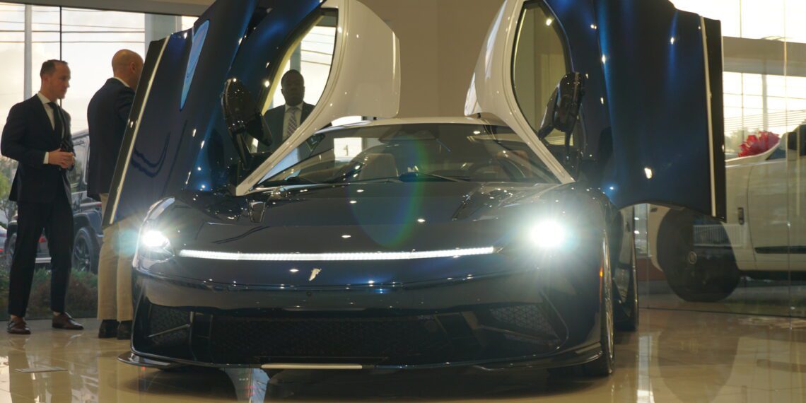 A luxury sports car with its doors open is displayed in Braman Motorcars' Palm Beach showroom. Nearby, three people stand conversing, perhaps captivated by the sleek design of Automobili Pininfarina.