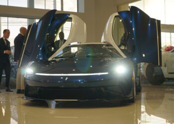 A luxury sports car with its doors open is displayed in Braman Motorcars' Palm Beach showroom. Nearby, three people stand conversing, perhaps captivated by the sleek design of Automobili Pininfarina.
