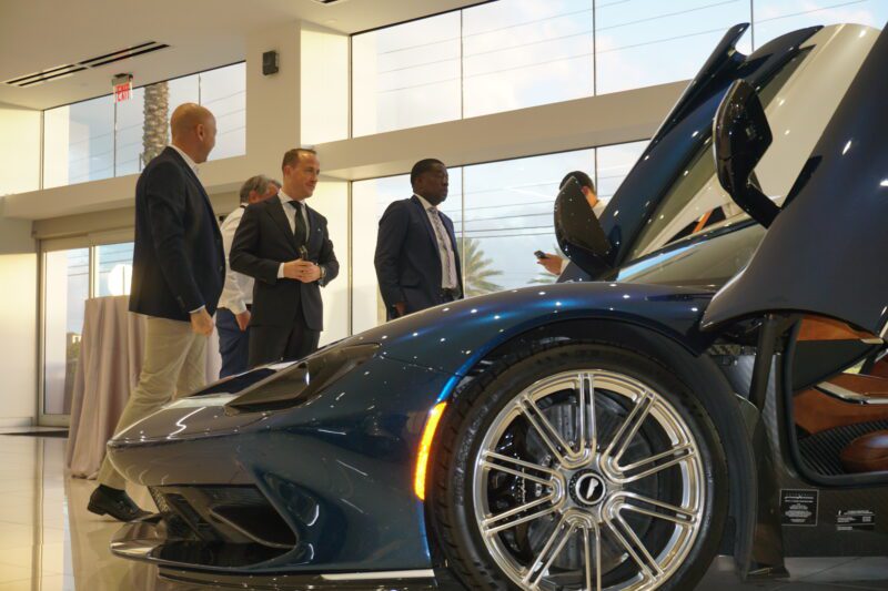 At Braman Motorcars in Palm Beach, a group of men in suits admires an Automobili Pininfarina blue sports car with open doors inside the showroom.