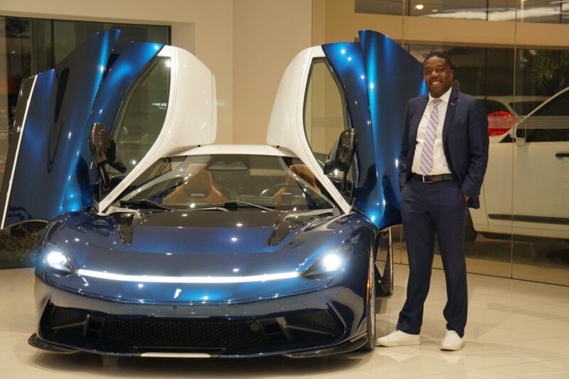 A person in a suit stands next to a blue sports car with gull-wing doors open at Braman Motorcars in Palm Beach, showcasing the elegance of Automobili Pininfarina's design.