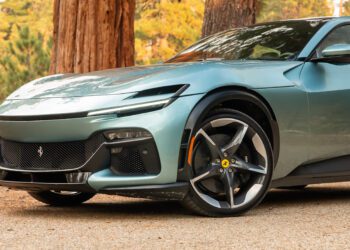 A sleek, light green V12 Grand Tourer sports car is parked on a dirt path, surrounded by tall trees. The iconic Prancing Horse emblem gleams proudly on its hood.