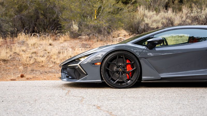 Close-up of the front side of a gray sports car parked on a road, showcasing a sleek design and red brake calipers. Dry grassy terrain and trees are visible in the background.