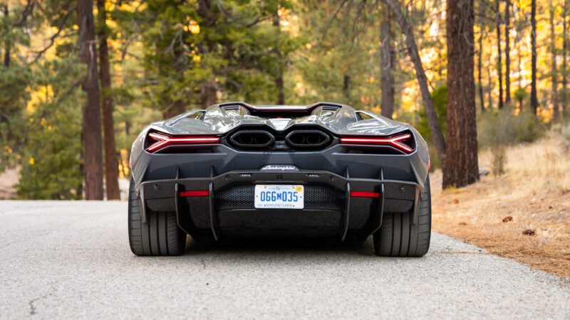 A black sports car on a forest road, viewed from the rear, with visible exhaust pipes and taillights.