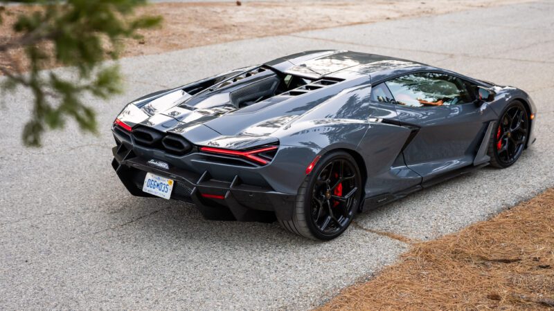 A sleek, grey supercar parked on a textured pavement, surrounded by natural scenery.