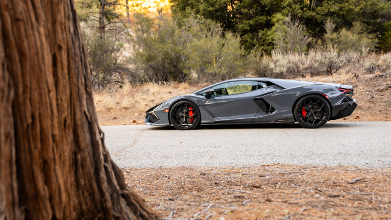 A sleek gray sports car on a paved road, surrounded by dry grass and trees in the background.