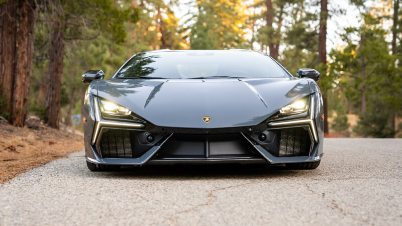 Front view of a sleek, black sports car parked on a forest road, surrounded by tall trees.