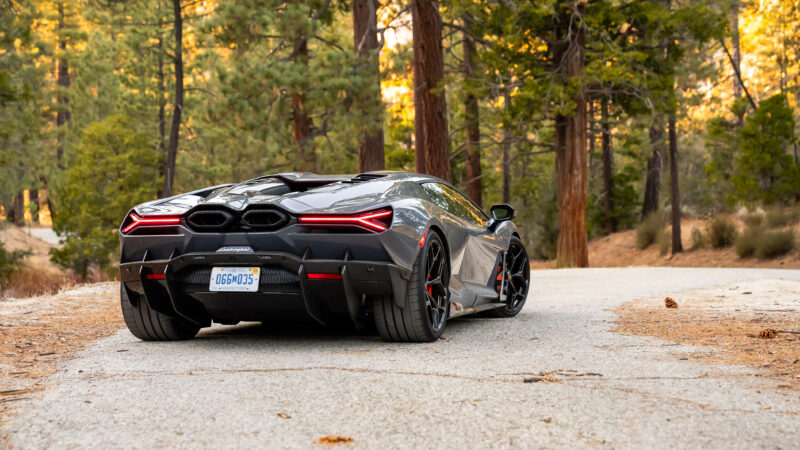 A sleek black sports car with red taillights is parked on a forest road surrounded by tall trees.