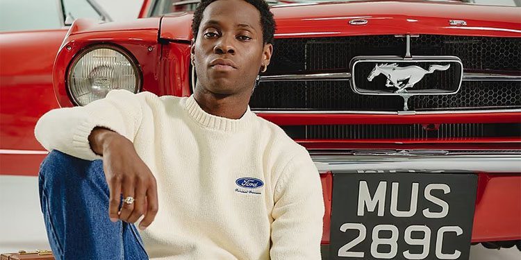 A person named Percival sits in front of a classic red Ford Mustang from the Ford Mustang Collection, proudly displaying its "MUS 289C" license plate, while wearing a white sweater and denim jeans as part of the 60th Anniversary celebration.