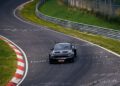 A black Ford Mustang GTD roars around the Nürburgring racetrack, surrounded by grass and guardrails. The track surface is marked with various writings and symbols, challenging drivers to push their limits under 7 minutes.