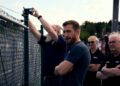 A group of men in dark shirts stand behind a chain-link fence, intently observing an outdoor scene. One man holds up a device, capturing moments as they eagerly await the Ford Mustang GTD aiming to conquer the Nürburgring in under 7 minutes.