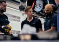 Three men in a garage workshop discuss something near a Ford Mustang GTD lifted on a hydraulic jack. One man gestures with his hands, perhaps talking about their last Nürburgring laps. A drill and tools are visible on the table.