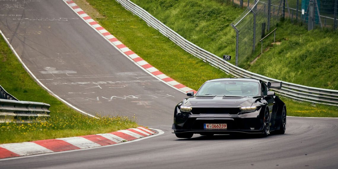 A black Ford Mustang GTD tears through the curvy, hilly racetrack of Nürburgring, surrounded by lush greenery.