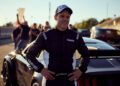 A race car driver in a black and white racing suit stands proudly in front of a Ford Mustang, with the iconic Nürburgring racetrack and an eager crowd buzzing in the background.