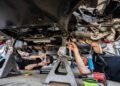 Two mechanics work under a raised Ford Mustang, using various tools to ensure it's ready for the demands of the Nürburgring. The car is supported by jack stands, each technician focused on their task amidst the hum of potential speed and precision.