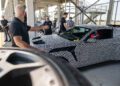 A group gathers near a camouflaged Ford Mustang GTD at the Nürburgring racetrack; one individual points while others observe intently. The car, poised to potentially conquer the track in under 7 minutes, is partially obscured by a blurred object in the foreground.