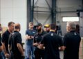 A group of eight men stand in a factory setting, some wearing black shirts and caps, engaged in discussion about the Ford Mustang GTD's impressive under 7 minutes lap at Nürburgring. Industrial equipment is visible in the background.