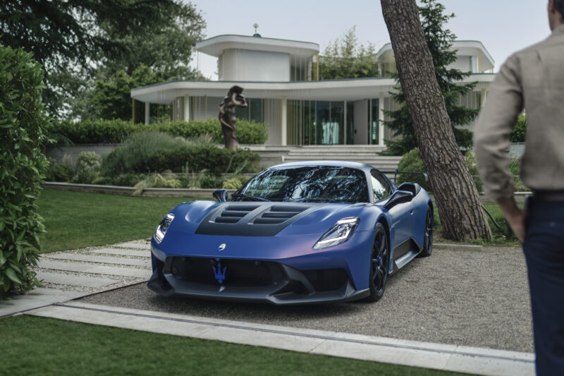 A blue sports car parked on a gravel driveway in front of a modern white house, surrounded by greenery and a statue.