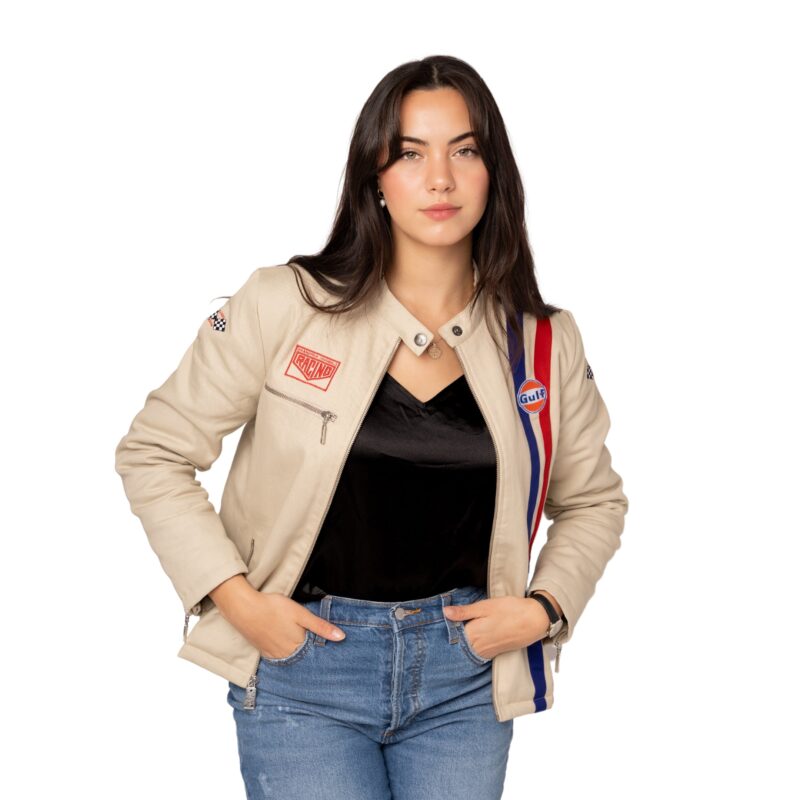 A person wearing a cream jacket adorned with patches, a black top, and blue jeans stands with hands in pockets against a white backdrop, embodying a stylish look perfect for the 2024 Holiday Gift Guide found in the latest duPont REGISTRY shop.