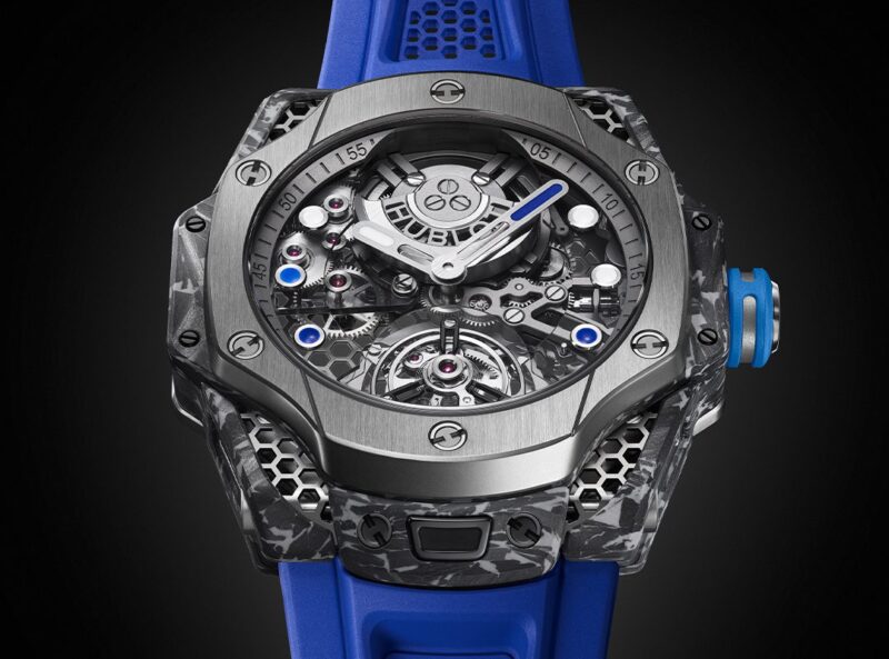 A detailed wristwatch, reminiscent of Hublot's innovative design, features a complex, exposed mechanical movement and a rugged gray case paired with a blue strap. This piece echoes the bold aesthetics of Samuel Ross' Big Bang Tourbillon collaboration.