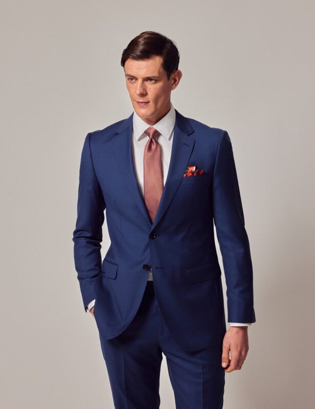 A distinguished gentleman in a blue suit and red tie stands elegantly against a plain background.