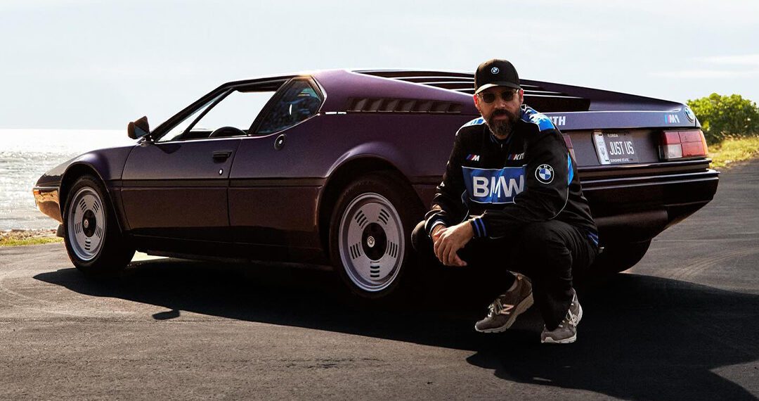 A man crouches beside a vintage purple sports car, reminiscent of Ronnie Fieg's collaborations, with custom license plate "MLS 16" on a sunny road by the sea.