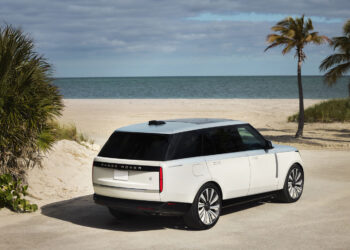 The white Range Rover SV Candeo is elegantly parked on a sandy beach with palm trees, overlooking a calm ocean under a cloudy sky.