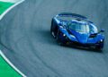 A Praga Bohema Supercar, valued at $1.5M, makes a sharp turn on a race track, with a section of green turf visible along the track edge.