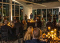 A formal gala unfolds in a dimly lit room in Middle Tennessee, with guests seated at elegantly set tables. Candles illuminate the scene as two speakers take center stage. Large windows reveal the twinkling city lights outside, creating a magical backdrop for the Make-A-Wish event.