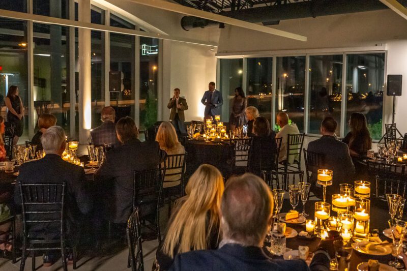 A formal gala unfolds in a dimly lit room in Middle Tennessee, with guests seated at elegantly set tables. Candles illuminate the scene as two speakers take center stage. Large windows reveal the twinkling city lights outside, creating a magical backdrop for the Make-A-Wish event.