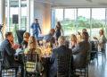 At the Middle Tennessee Gala, people seated at round tables in a room with large windows listen to a man and woman speaking at the front. Candles adorn the tables, and greenery is visible outside, creating an enchanting ambiance where dreams come true.