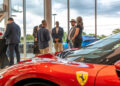 People are gathered in a car showroom for the Make-A-Wish Gala, standing near a red Ferrari with its logo visible. Large windows in the background reveal an overcast sky and lush greenery, setting the perfect scene in Middle Tennessee.