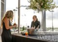 A woman in a black dress chats with a bartender at a table adorned with a bucket of ice and bottles. Glasses are neatly arranged under a large floral arrangement near sunlit windows, setting the scene for the elegant Make-A-Wish Gala in Middle Tennessee.