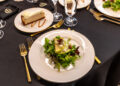 A plate of mixed greens with apple slices and cheese crumbles sits next to a slice of cheesecake, elegantly arranged on a table adorned with gold utensils, capturing the essence of the Middle Tennessee Gala where dreams come true.