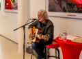 At the Middle Tennessee Gala, a gray-haired musician strums an acoustic guitar, seated beside a red table adorned with a water bottle and papers. This touching performance celebrates the power of making dreams come true.