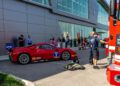 A stunning red race car is parked outside a modern building, drawing a curious crowd. Among them are two individuals in firefighting uniforms, adding an air of excitement reminiscent of a Middle Tennessee gala. In the background, a fire truck is partially visible.