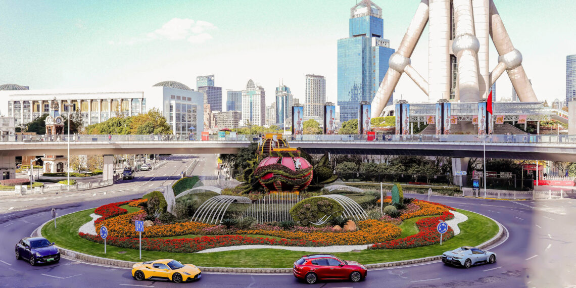 A roundabout with a floral display, surrounded by Italian luxury cars like Maserati, graces the urban setting. Modern buildings frame the scene, while a large tower in the background adds to the sophisticated ambiance perfect for celebrating 110 years of elegance and innovation.