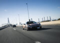 A sleek, light brown Maserati sports car effortlessly cruises down the highway, leading a parade of vehicles. Celebrating 110 years of Italian luxury, it glides smoothly with a bridge visible in the distance under a clear blue sky.