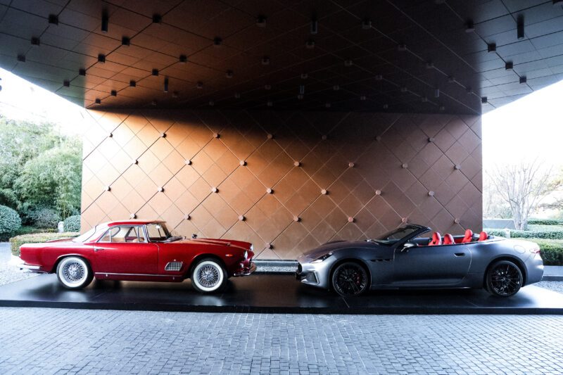 A vintage red coupe and a modern gray convertible, both embodying Italian luxury, are displayed side by side on a platform with a patterned background.